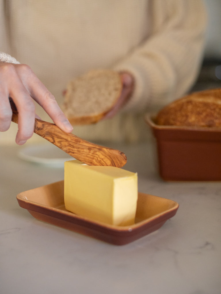 Someone scrapping some butter to put on a slice of bread beside a pan of bread in a loaf pan. 