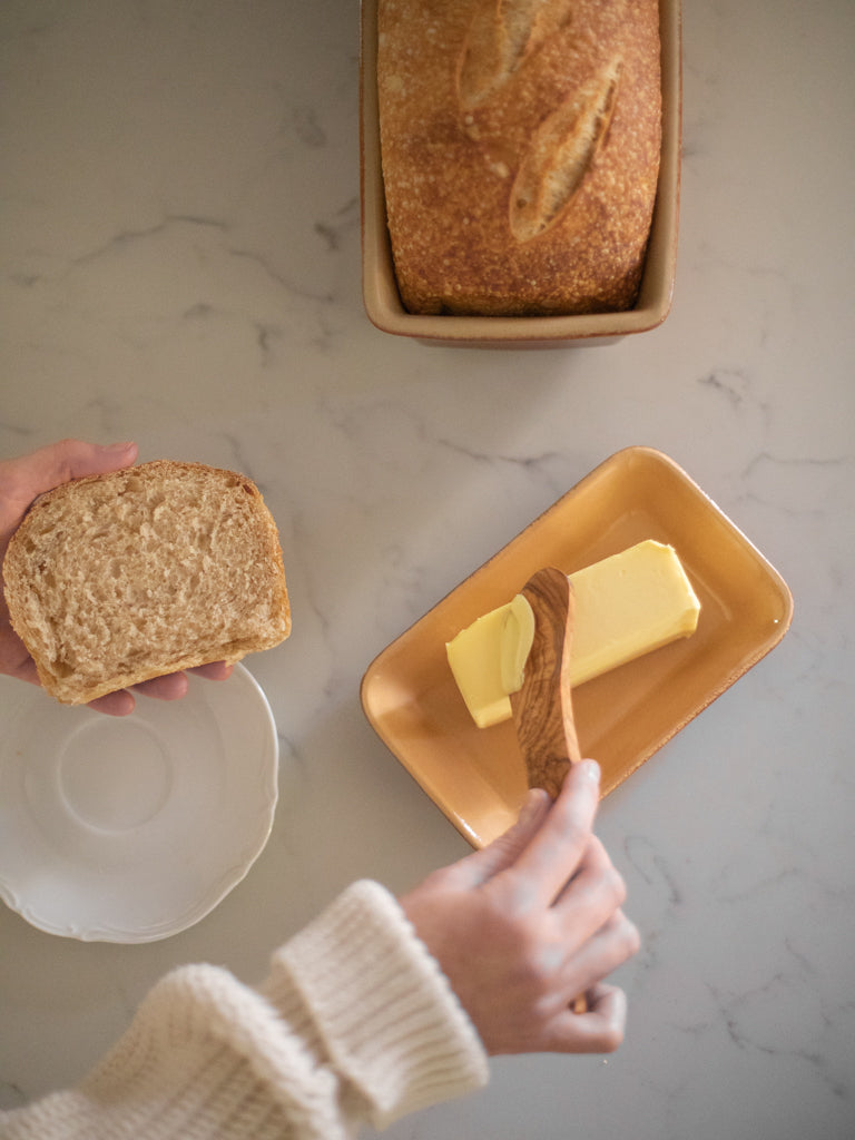 Someone scrapping some butter to put on a slice of bread beside a pan of bread in a loaf pan. 