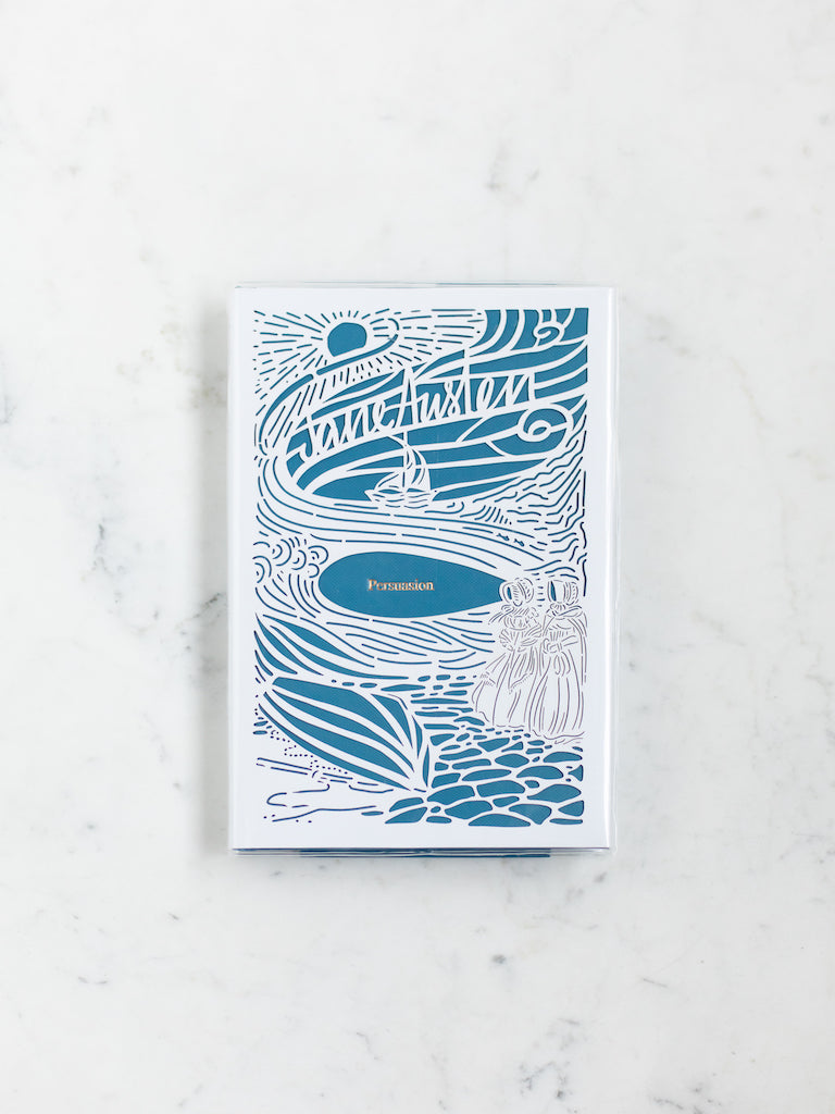 A blue book with a white paper cut design over it of two women in bonnets on a beach with a ship in the distance. 