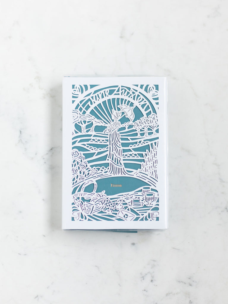 A blue book with a white paper cut design over it of a woman with a parasol standing behind a picnic blanket with trees behind her. 