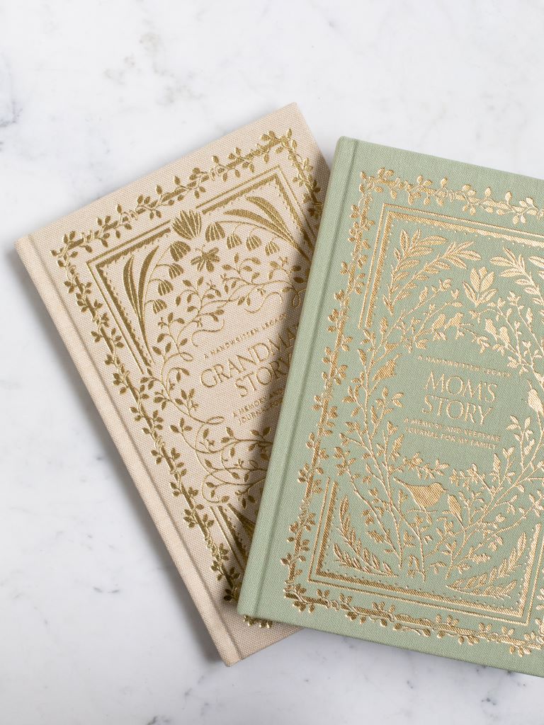 A green clothbound book with gold designs of birds and plants around the title “Mom’s Story” with a cream clothbound book with gold designs of plants and flowers around the title “Grandma’s Story” under it. 