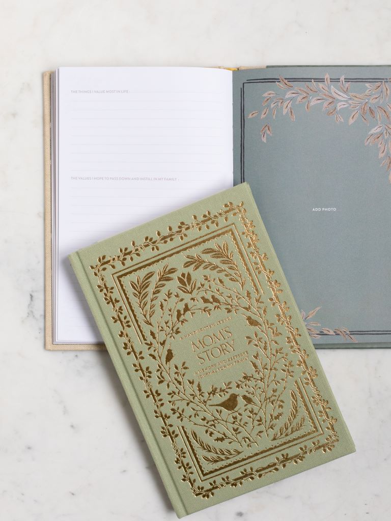 A green clothbound book with gold designs of birds and leaves around the title “Mom’s Story” with an opened copy of the book behind it. 