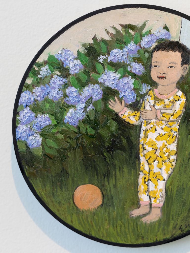 A small circular canvas displaying a painting of a small child in a yellow and white onesie, standing with its arms out in a grass field next to a bush of purple flowers, with a small orange ball on the ground.