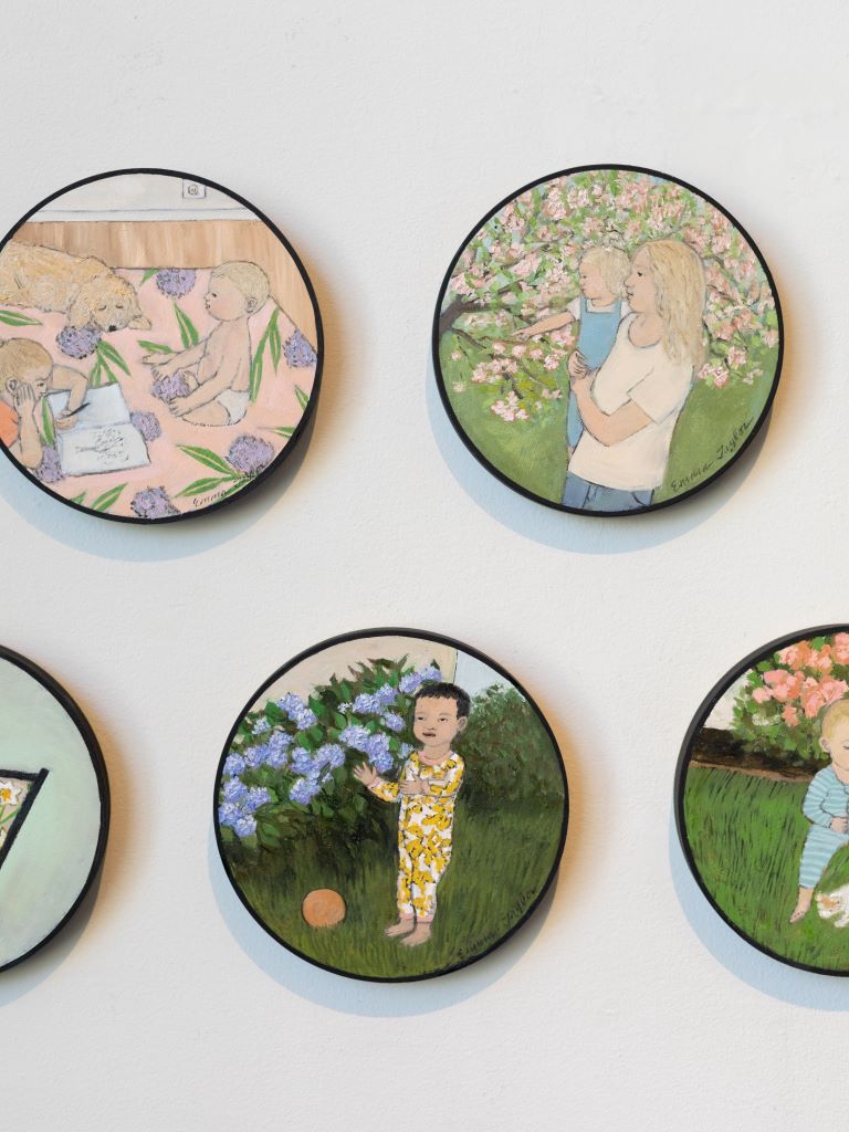 A small circular canvas displaying a painting of a small child in a yellow and white onesie, standing with its arms out in a grass field next to a bush of purple flowers, with a small orange ball on the ground. It is hung on a wall among other painting in the same frame depicting different settings and scenarios with children.