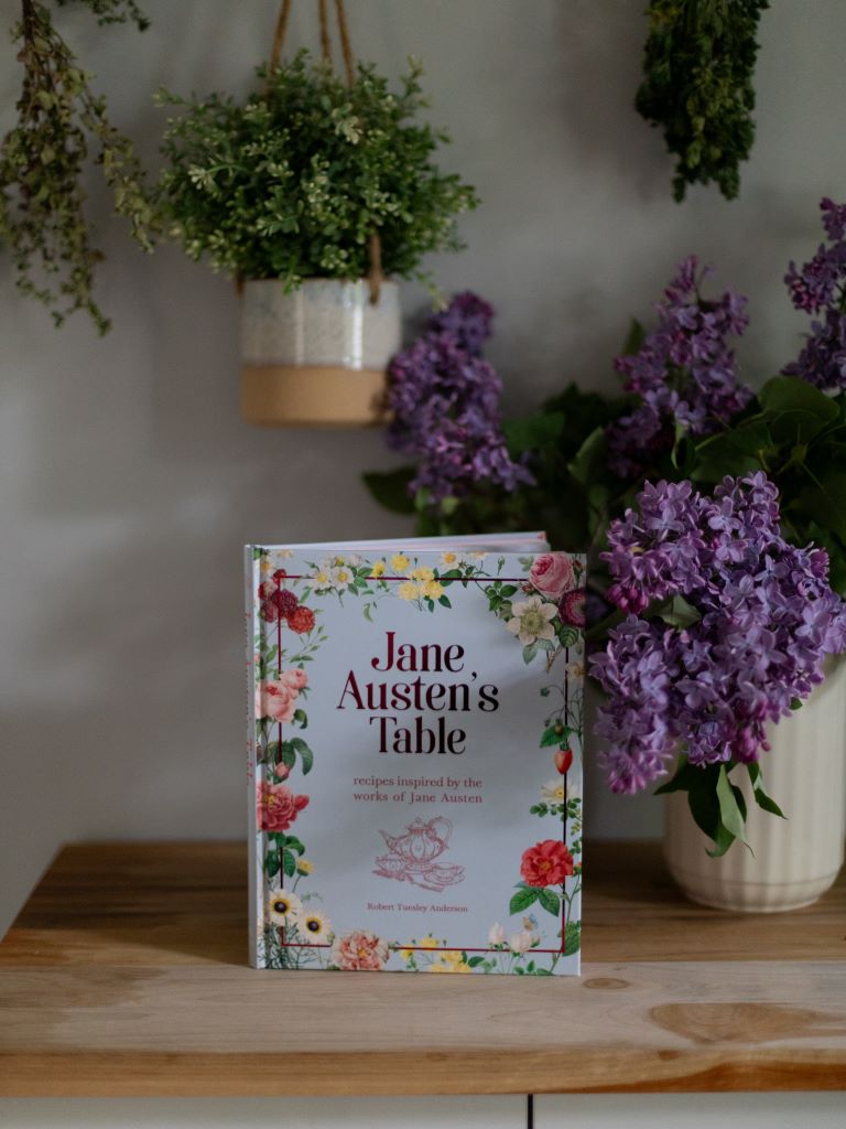 A blue cookbook covered in flowers on a table beside some lilacs in a vase. 