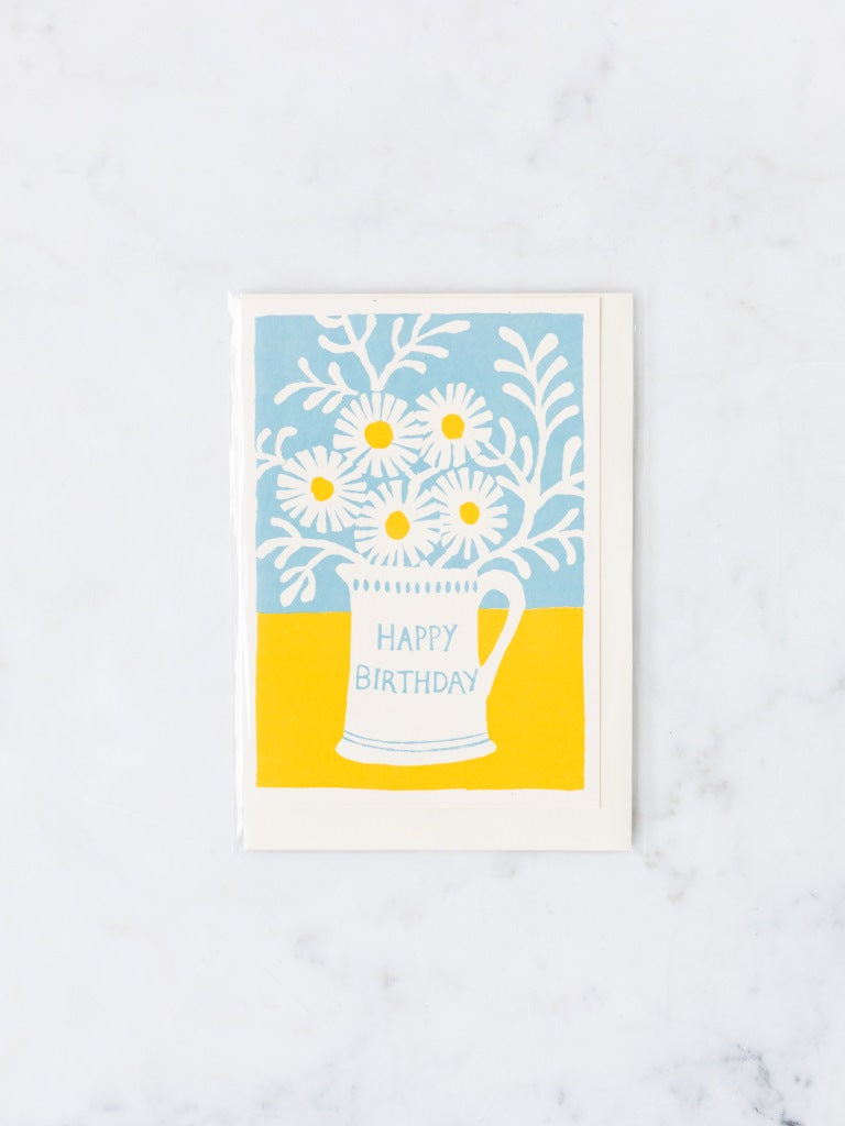 Blue and yellow colors with a pitcher holding a jar of white daisies happy birthday card.