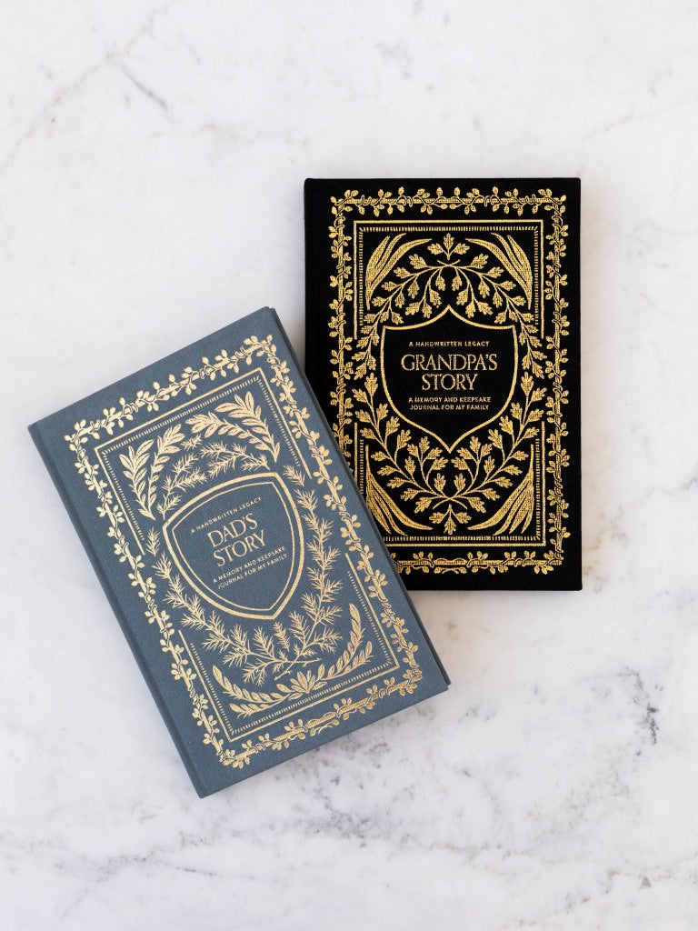 A blue clothbound book and a black clothbound book with gold designs of plants surrounding their titles. The blue one with the title “Dad’s Story” the black one with the title “Grandpa’s Story.” 