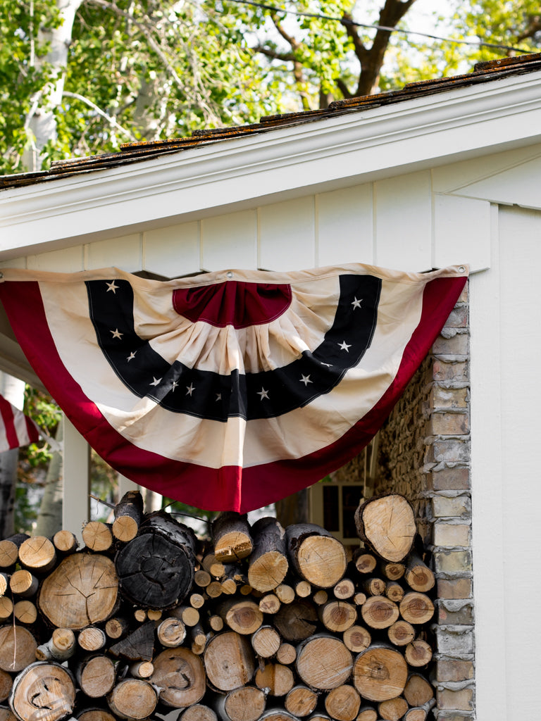 Photo of Americans bunting flag hanging on a shed over a pile of logs. The bunting has natural creases and lays relaxed down the side of the shed.