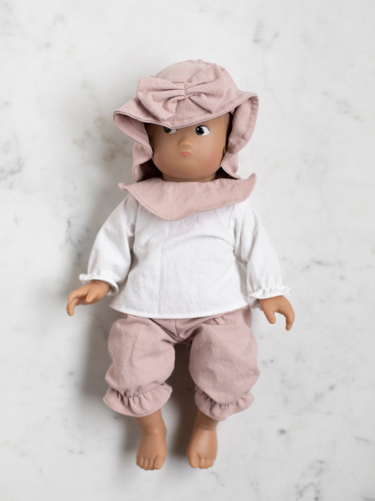The baby doll lays flat in its back and is wearing a pink bow hat with pink pants, a white shirt, and a pink bib. The baby doll has an olive skin tone.