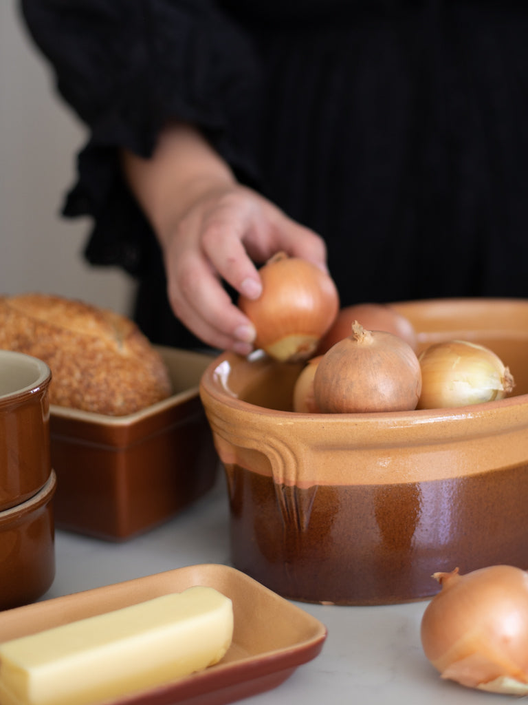 A person putting onions into a dish next to a pan with bread inside and a dish of butter. 