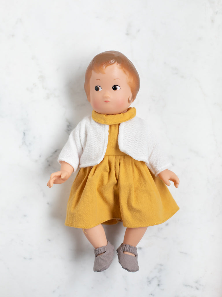 The baby doll lays flat on its back with a mustard dress, white sweater, and brown slippers. The baby doll has ginger hair coloring and light skin tone.