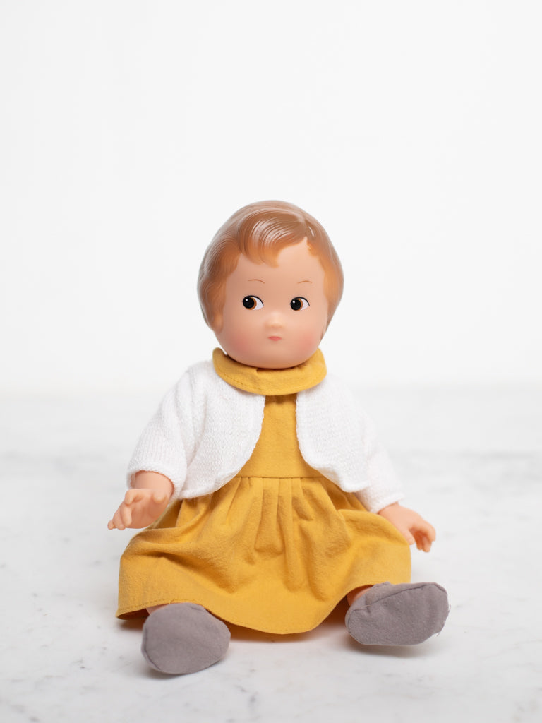 A baby doll sits front view with a mustard yellow dress, a white sweater, grey slippers, and ginger hair.