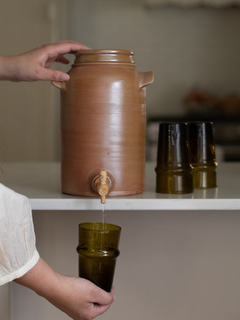 Someone using a tall brown water jug to fill up a glass. 
