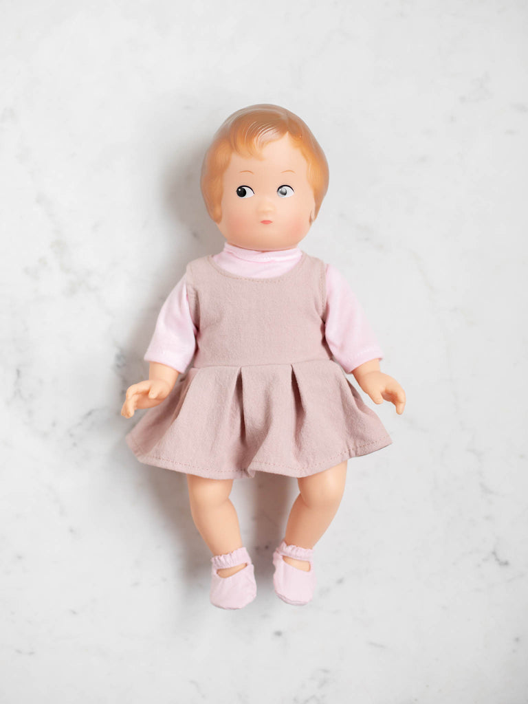 A baby doll lays flat on their back with a baby pink shirt, plush dress, and baby pink slippers. The baby doll has ginger hair coloring and a light skin tone.