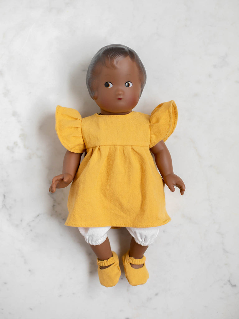 A baby doll with a mustard dress, white stocking, and mustard slippers. This baby doll has dark brown hair and a darker skin tone. 