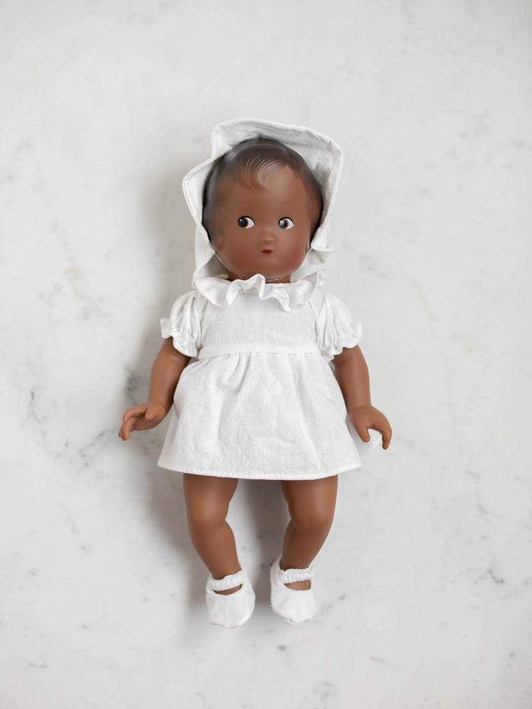 The baby doll lays on its back with front view. The doll has a delicate white bonnet, a white dress, and white slippers. The doll also has dark brown hair and a dark skin tone.