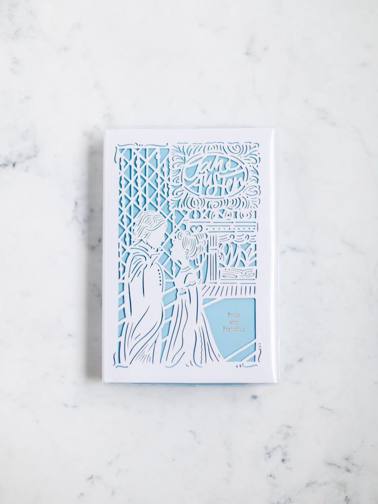 A blue book with a white paper cut design over it of a man and a woman looking at each other by a fire in a living room. 