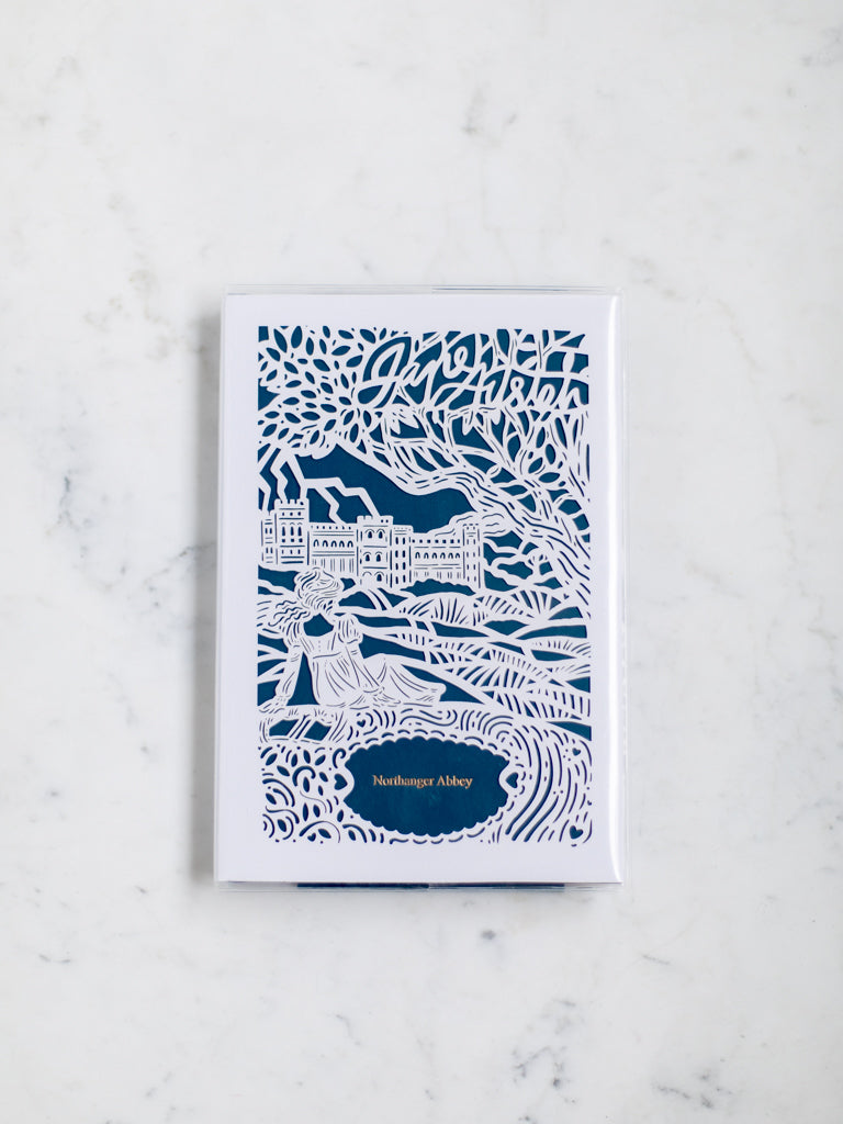A blue book with a white paper cut design over it of a woman lying under a tree  gazing at a distant mansion. 
