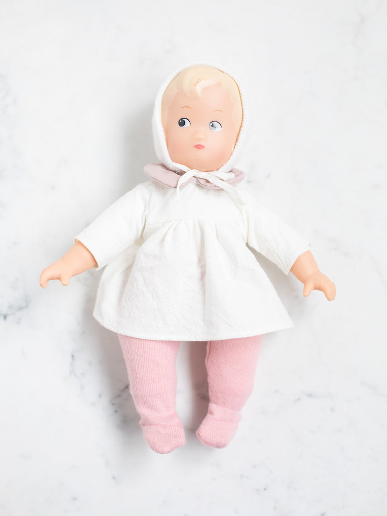 The baby doll lays flat on its back with pink pants, a white dress with a white hood and pink bow. This baby doll has blonde hair and a peach skin tone. 