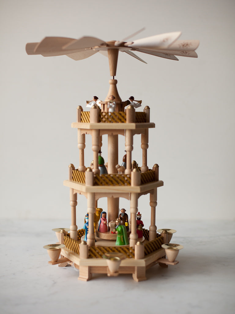 A large spinning wooden three tiered decorative structure that gradually gets smaller toward the top. Contains multiple characters representing those of the Nativity scene. A large windmill-like spinning panels facing upwards on top. 