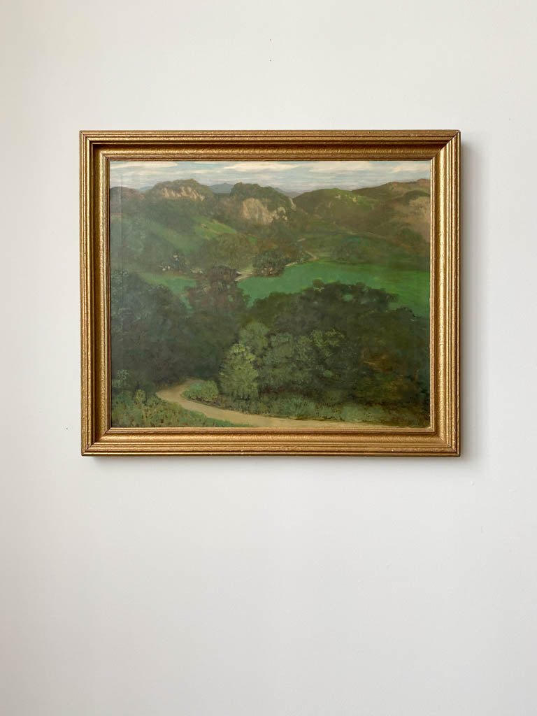 Painting of a trail through some trees to a canyon in a gold frame hung on a wall. 