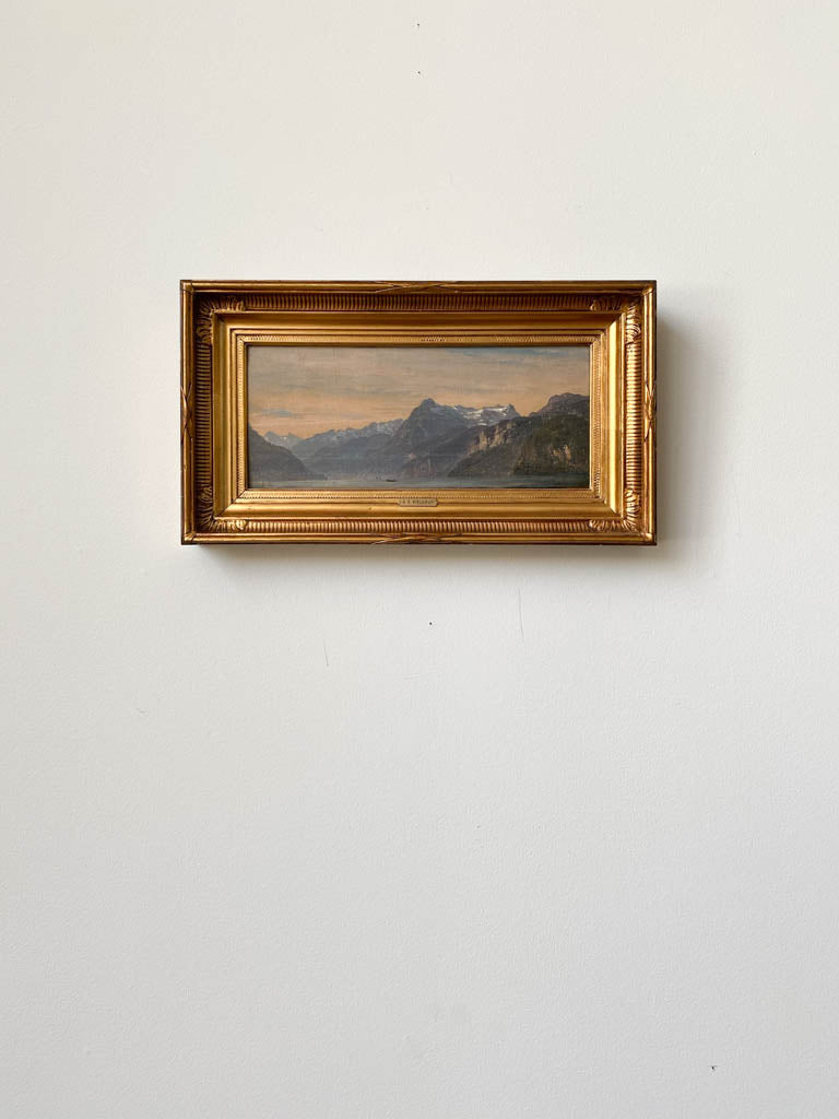 A painting of a boat in a lake beside some huge mountains hung in a gold frame. 