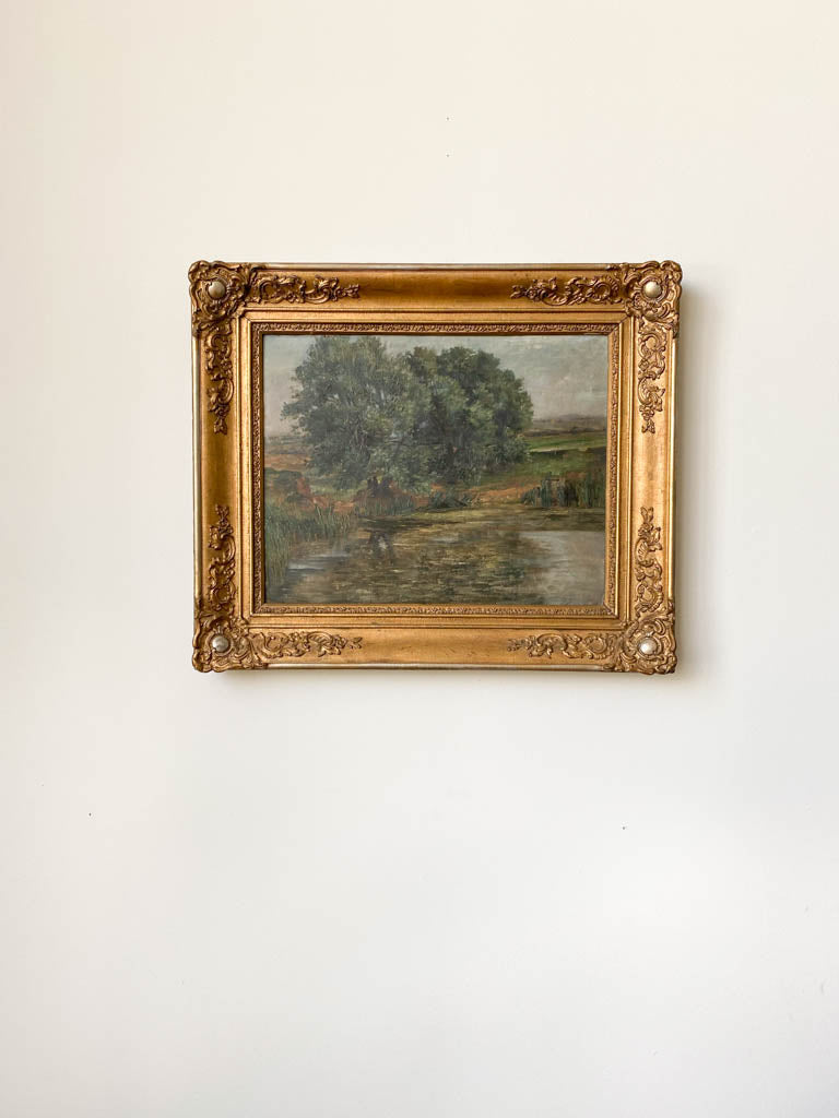 Painting of a pond by a tree in a gold frame on the wall. 
