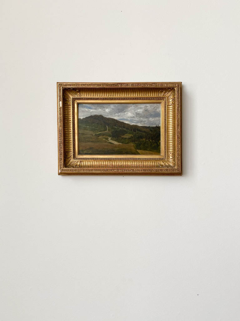 A painting of a green hilly landscape in a gold frame on the wall.  