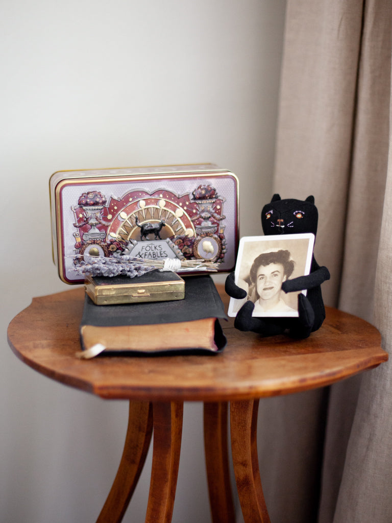 A table with some old heirloom books, a stuffed cat holding up a photo of a relative, and the Folks and Fables tin on top of it. 