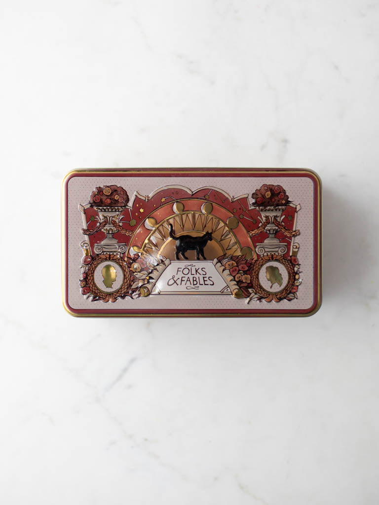 Folks and Fables tin with orange and gold colors featuring a cat and profile faces beside flower vases. 