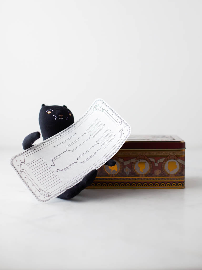 A stuffed cat holding a family tree beside the Folks and Fables tin. 