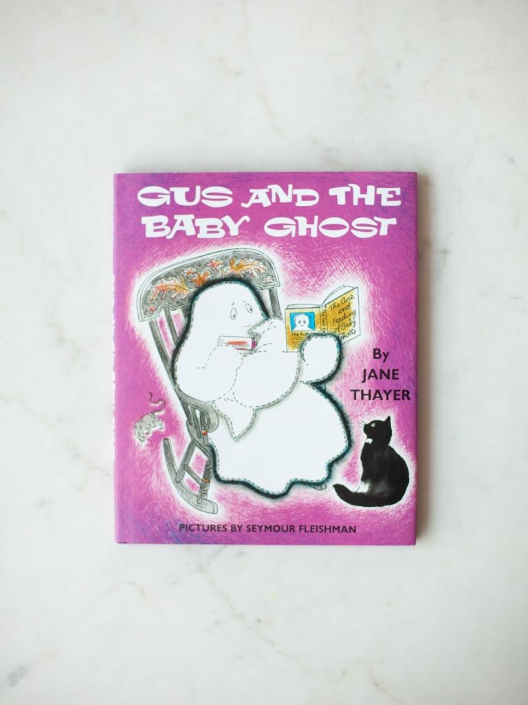 Gus and the Baby Ghost book cover of Gus sitting in a rocking chair while holding a baby blanket and feeding it a bottle, while also read it a book. A black cat looks up from the floor.