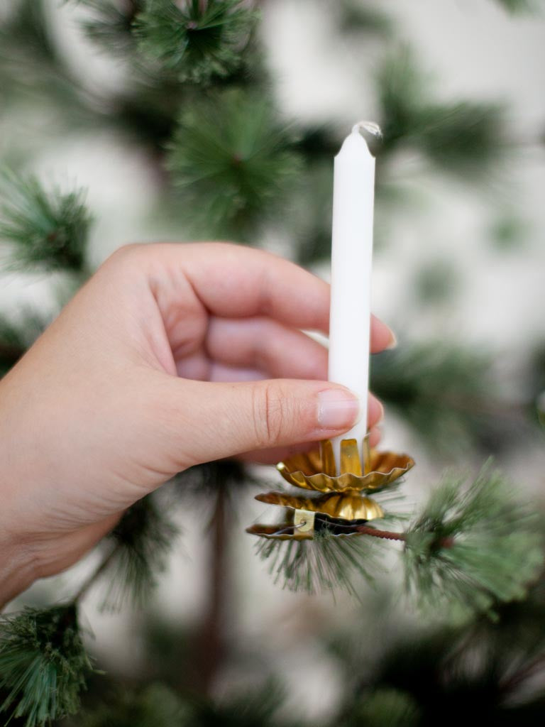 A piece of gold metal in the shape of an open flower, with a space to hold a candle in the middle all sitting on top of a clip. A white candle in the middle and held onto by a hand as it is being placed on the branch of a Christmas tree.