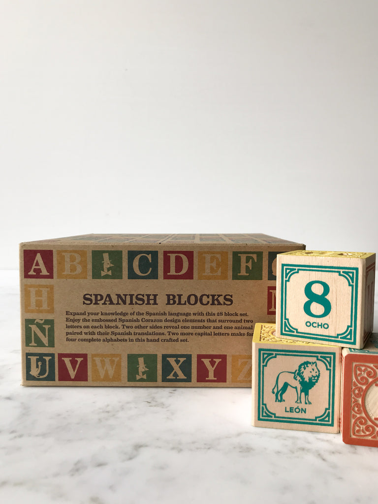 Some blocks decorated with letters, numbers, and animals on their sides with the box they come in behind them titled “Spanish Blocks.” 