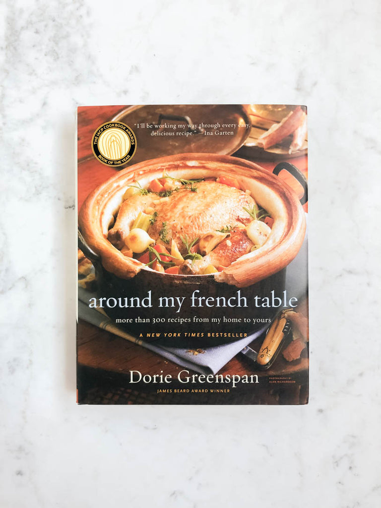 Front view of a cookbook entitled “around my French table”. The cover of the book surrounds a chicken roasted in a Dutch oven with various herbs and vegetables.