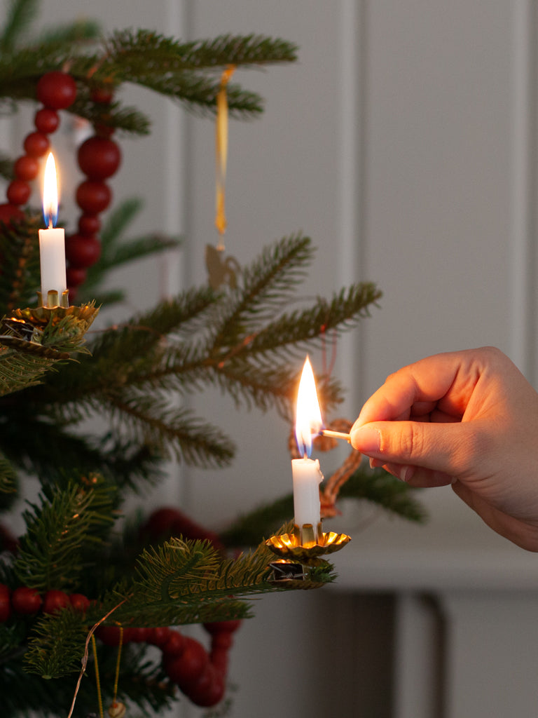 A piece of gold metal in the shape of an open flower, with a space to hold a candle in the middle all sitting on top of a clip. A white candle in the middle being lit as it sits on the branch of a Christmas tree.