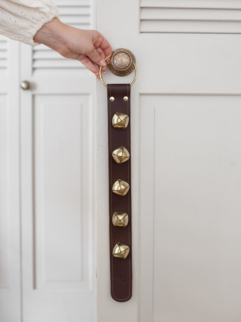 Someone hanging a leather strap with sleigh bells on it on a doorknob by a gold hoop at the top. 